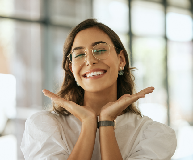 Smiling woman wearing glasses and white shirt in bright office setting, hands raised expressively.