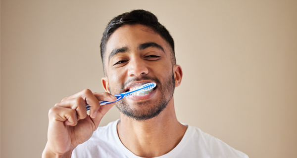 Man brushing teeth with blue toothbrush against neutral beige background, smiling.
