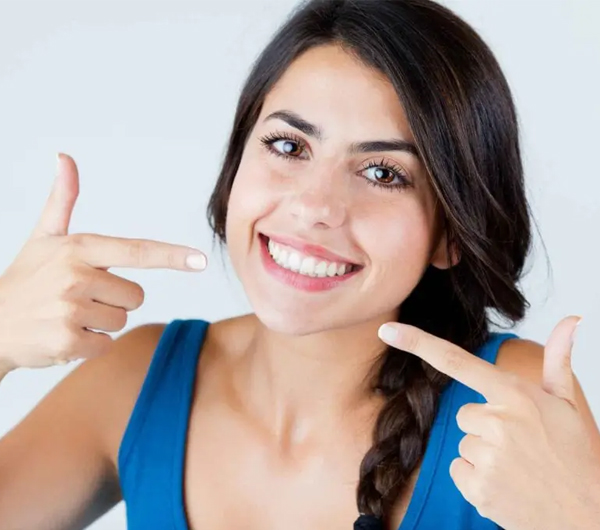 Young woman smiling brightly, pointing at her teeth with both hands against white background.