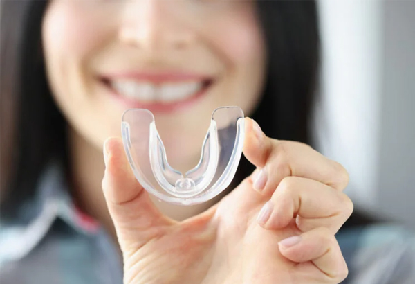 Woman smiling while holding clear dental retainer or aligners in her hand indoors.