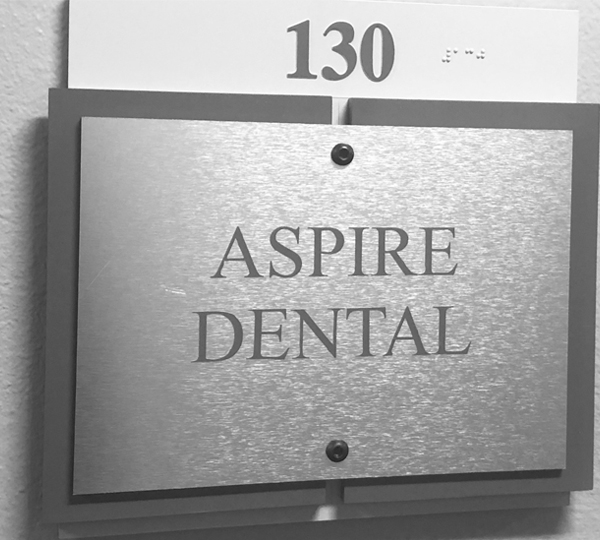 Brushed metal office sign reading "ASPIRE DENTAL" with room number 130 mounted on wall.