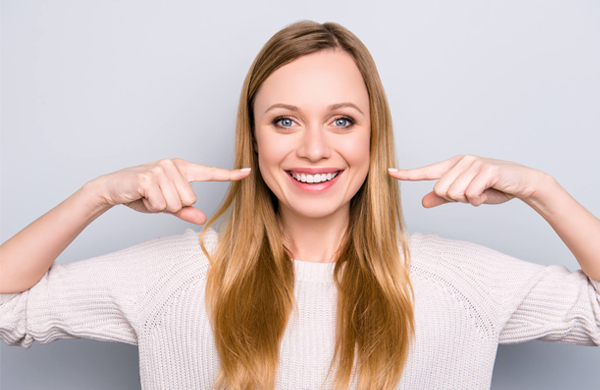 Smiling woman with long hair pointing to her teeth against neutral background.