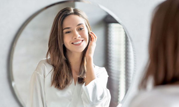 Woman smiling at reflection in round mirror, wearing white shirt in dental office setting.