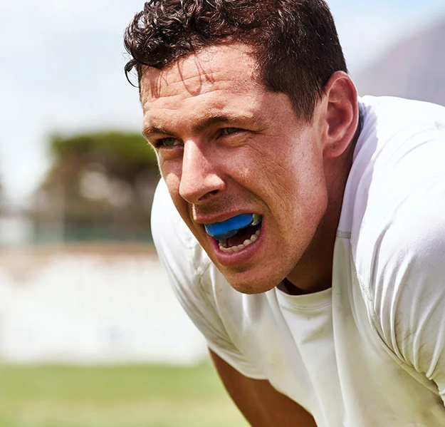 Male athlete wearing blue mouthguard in white shirt on grass field outdoors.