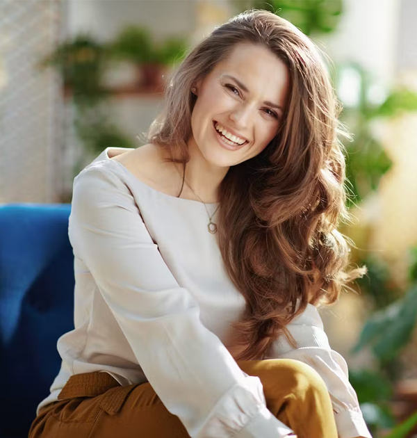 Smiling woman with long brown hair wearing white blouse in dental office setting with plants.