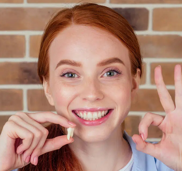 Young woman with red hair smiling broadly, pointing to her teeth against brick wall background.