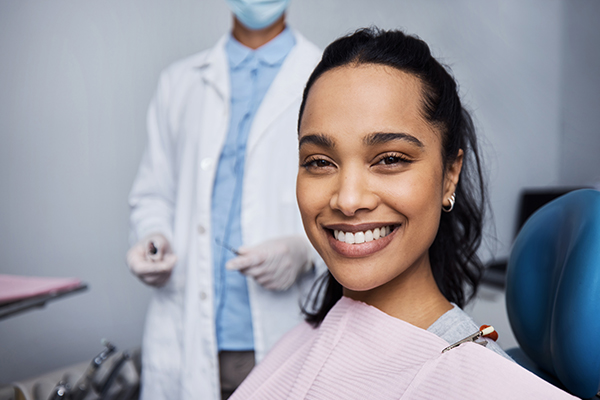 Smiling patient in dental chair with dentist in white coat in background at clinic