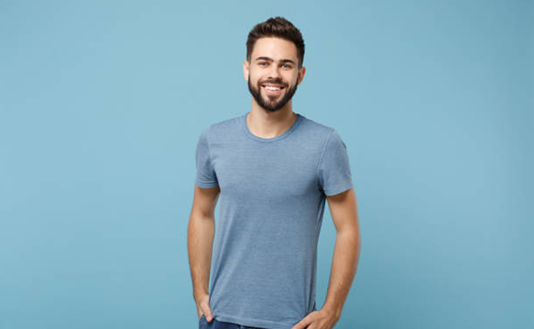 Man in blue t-shirt standing against light blue background, smiling at camera.