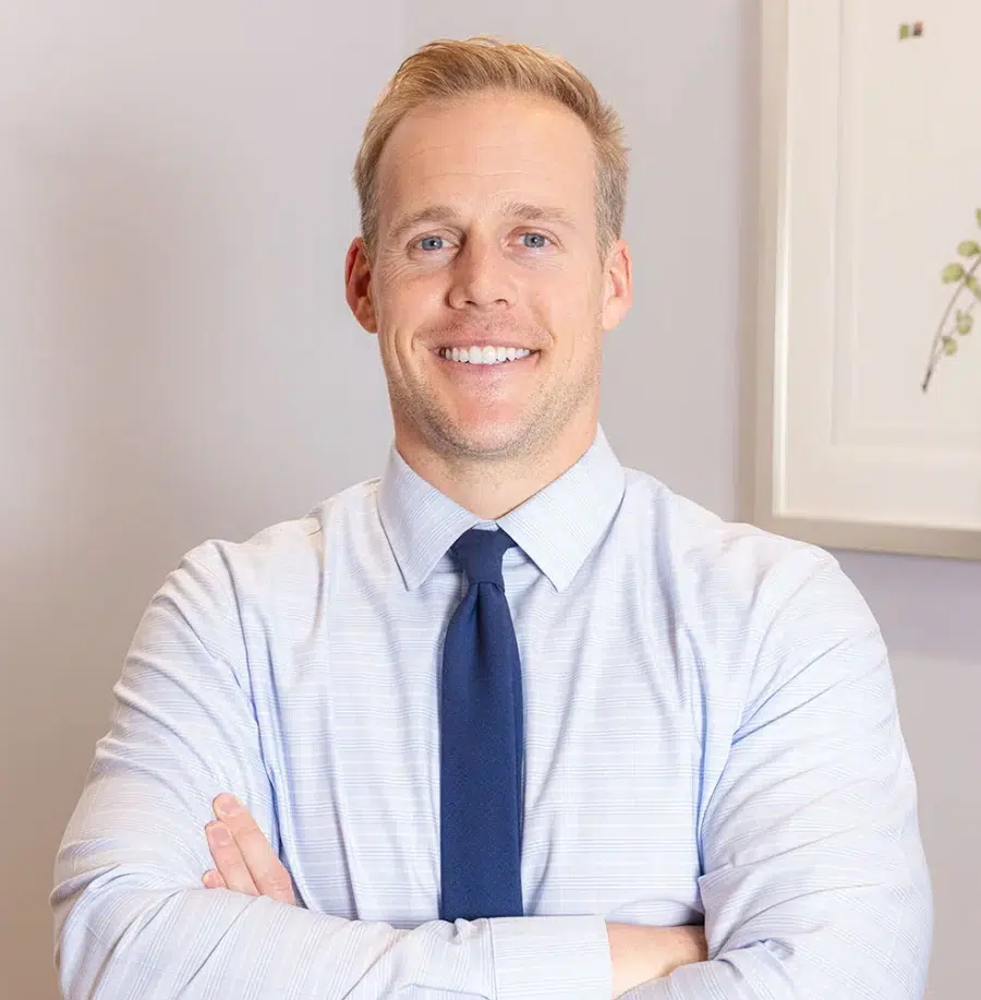 Professional male dentist in light blue shirt and navy tie, arms crossed, smiling in dental office setting.