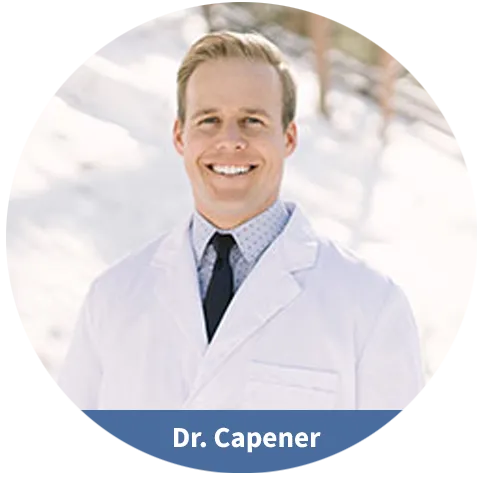 Dr. Capener wearing white lab coat and tie, smiling at camera in dental office setting.