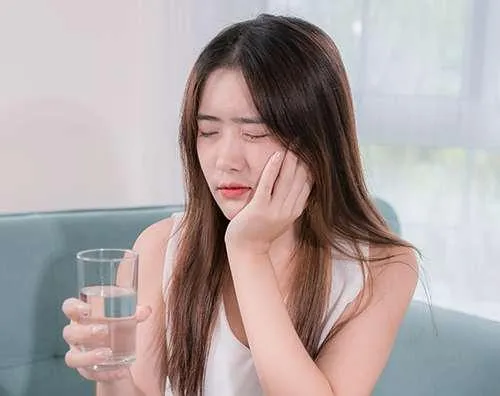 Woman holding water glass looking unwell, touching face in modern dental office setting.