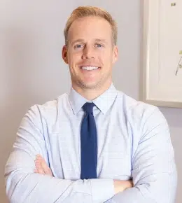 Professional male dentist in light blue shirt and navy tie, smiling with arms crossed in dental office setting.