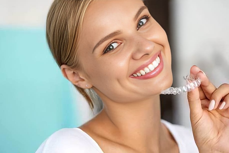Woman smiling while holding clear dental aligner in bright dental office setting.