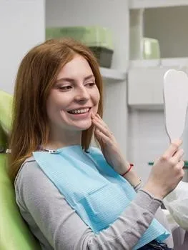 Woman in dental bib examines her smile in mirror at dental office.
