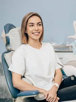 Smiling woman in white shirt seated in modern dental chair in bright office setting.